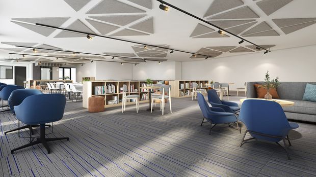 A bright, open-plan commercial lounge and library area with blue upholstered lounge chairs, small round tables, communal tables, low bookcases filled with books, and a grey patterned carpet. White walls and ceiling panels feature geometric acoustic treatment, with black track lighting throughout. Glazed doors and windows open to adjacent work or meeting areas.