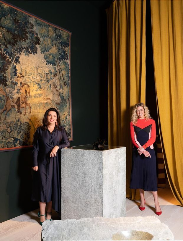 A dramatic, gallery-like interior featuring two women standing beside a large textured stone or concrete pedestal. The room has deep charcoal walls, a large traditional tapestry artwork, rich mustard-gold theatrical curtains, pale timber flooring, and sculptural decorative objects. The styling is highly curated and theatrical with strong contrast between dark walls, golden drapery, and vivid clothing.
