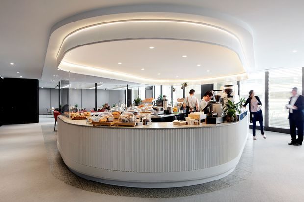 A bright contemporary commercial hospitality space centred on a large rounded, oval island counter displaying food and beverages. The counter has ribbed white fixed joinery with a dark benchtop, surrounded by tables, plants, glass partitions, and people. A sculptural recessed ceiling with layered curved edges and integrated linear lighting defines the space, with large windows and horizontal blinds along the right side.