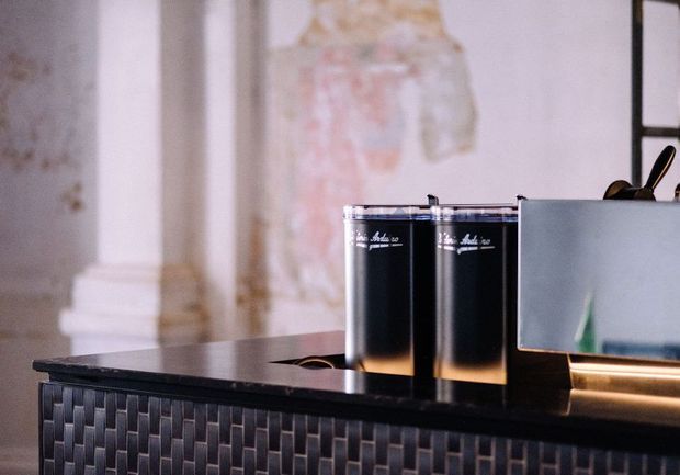 Close-up of a refined bar or reception counter featuring a dark tiled front with a repeating rectangular relief pattern, a glossy dark benchtop, metallic trim, and two black beverage cans. The background shows ornate pale architectural detailing and a framed artwork, giving the setting a sophisticated hospitality character.
