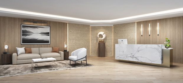 A refined contemporary reception or living space with a warm neutral palette. Vertical timber slat wall panels frame a textured stone feature wall and artwork, while a veined marble reception counter anchors the right side. The room includes a beige sofa, sculptural lounge chair, low coffee table, wood-look flooring, integrated linear lighting and understated decorative elements.