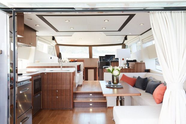Bright, refined interior of a modern luxury motor yacht or similar compact marine accommodation, combining a galley kitchen with integrated appliances, timber cabinetry, a raised dining area, and an upholstered lounge. Large windows and drawn white curtains create an airy atmosphere, while dark trim outlines the recessed ceiling.