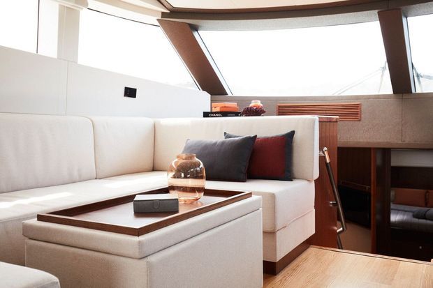 Bright, refined living area aboard a modern vessel, featuring a large cream upholstered sectional sofa, dark accent cushions, a low square coffee table with a wood-toned trim, and warm timber flooring. Large angled glazing and exposed structural framing create an airy, contemporary setting, with built-in cabinetry and a stair or passage opening visible to the right.