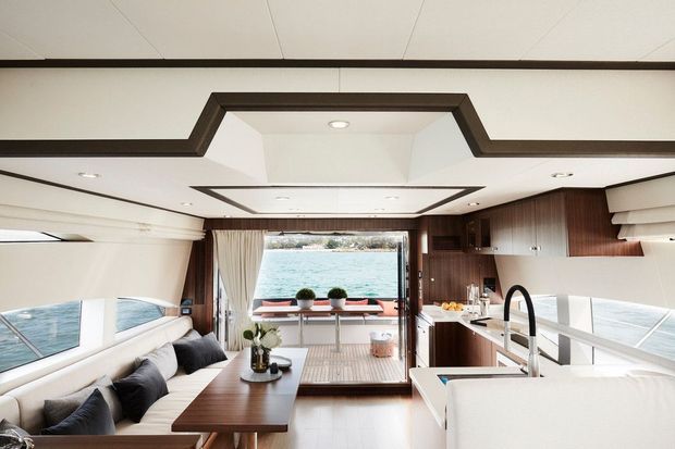 Bright, refined yacht interior combining a compact galley kitchen with an integrated dining and lounge area. Dark timber cabinetry and trim contrast with white upholstered seating, white surfaces, and a sculptural stepped ceiling. Large side windows and rear glazing provide expansive views over the water.