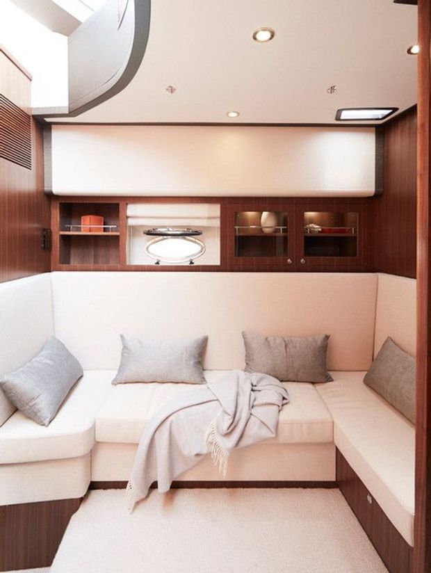Compact, refined lounge interior with a cream U-shaped built-in upholstered seating arrangement, pale cushions and throw, dark wood wall cabinetry and display shelving, and integrated recessed lighting. A curved ceiling detail and rounded built-in forms give the space a streamlined, likely marine-inspired character.