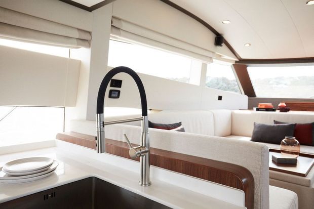 Bright, refined contemporary yacht interior showing a compact galley kitchen in the foreground with a white countertop, undermount sink and tall black-and-chrome gooseneck faucet. Beyond it is a built-in cream upholstered lounge with dark accent cushions, warm timber trim, integrated blinds, recessed ceiling lights and large windows.