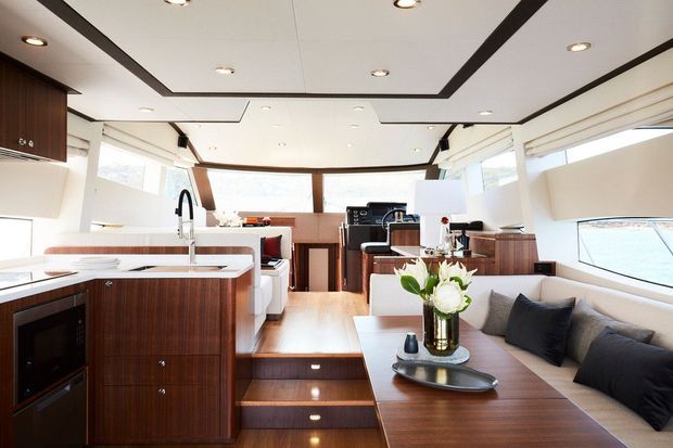 Bright, refined open-plan yacht interior with a compact galley kitchen, dining and lounge seating, warm timber cabinetry and flooring, white ceiling panels with dark recessed trim, and expansive wraparound glazing. A stairway links the raised aft seating area with the lower galley level.