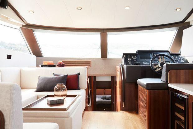 Bright contemporary boat cockpit and lounge interior with a white upholstered seating area, wood-trimmed cabinetry, a central helm with steering wheel and instrumentation, broad angled glazing, and a light paneled ceiling. The space has a clean nautical design with warm timber accents and abundant daylight.
