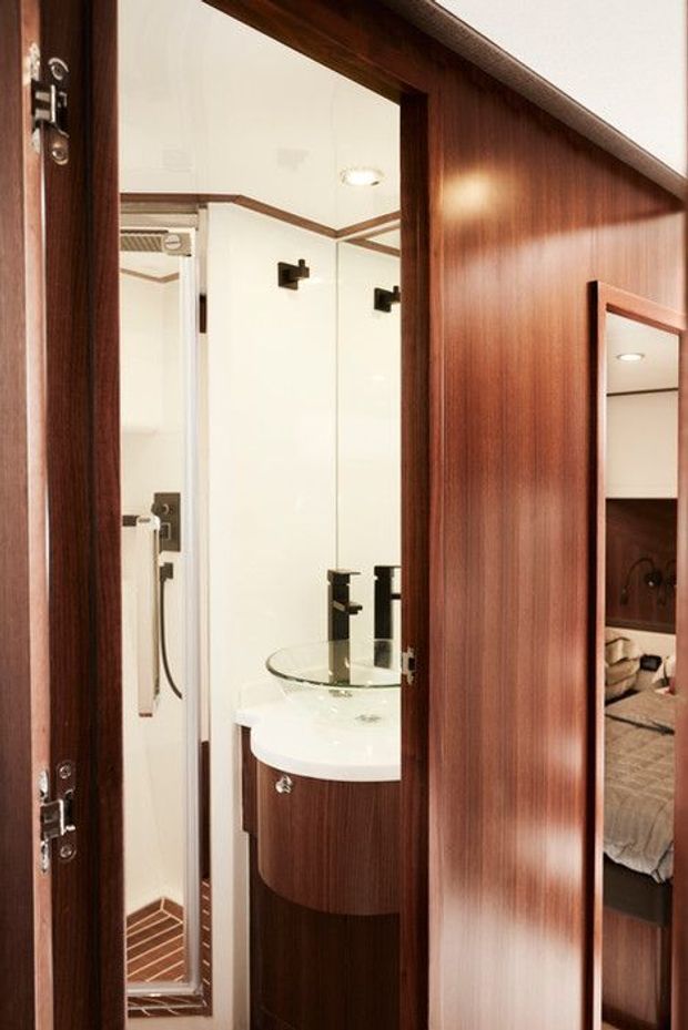 A compact, refined bathroom viewed through a partially open dark timber door. The room includes a curved white vanity with a glass vessel basin, dark fixtures, a mirrored wall, and a narrow tiled shower enclosure. Rich reddish-brown timber wall panelling and joinery dominate the surrounding space, with a partial bedroom visible through a second opening and reflected in the mirror.