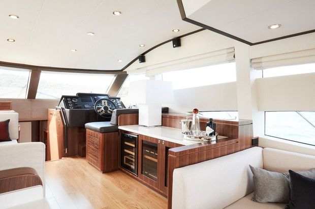 Bright, refined yacht interior with a compact galley and integrated bar cabinetry, navigation controls, light timber flooring, extensive windows, and built-in upholstered seating. The space has white ceiling and walls, warm wood joinery, dark trim, and abundant natural light.