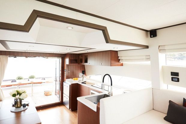 Bright open-plan galley kitchen and living area with warm dark timber cabinetry, white benchtops and splashback, integrated sink with a black gooseneck tap, and a white built-in sofa or banquette. A timber-framed glazed door opens to an outdoor deck, while a distinctive stepped ceiling bulkhead defines the space.