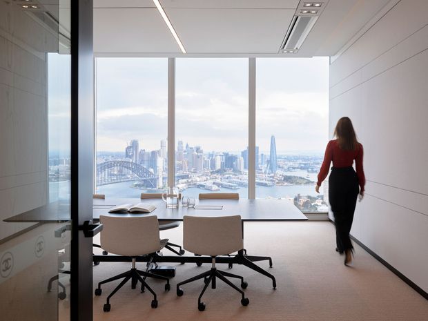 A refined contemporary office or meeting room with a long minimalist conference table, rolling upholstered chairs, integrated linear ceiling lighting, and floor-to-ceiling glazing. The room overlooks a prominent urban waterfront skyline with a large suspension bridge and high-rise buildings. A glass wall or door and sleek built-in surfaces reinforce the corporate architectural setting.
