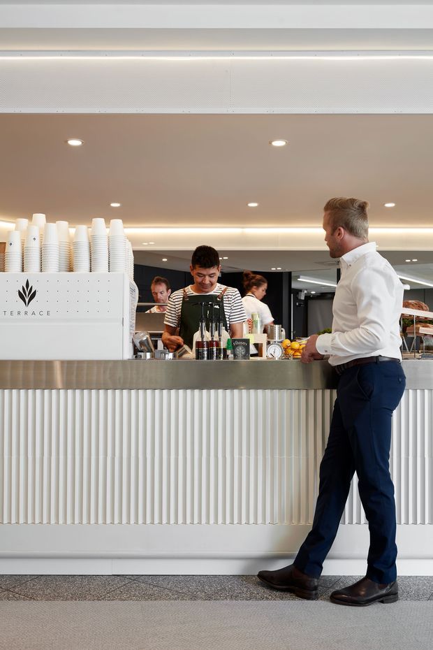 A bright, contemporary café or hospitality bar interior with a long white counter featuring vertically fluted panel cladding and a brushed metal benchtop. Baristas work behind the counter, with stacked takeaway cups, coffee equipment, bottles, fruit, and a visible Terrace logo. Recessed downlights and concealed linear lighting illuminate the warm neutral space, while a customer stands at the counter in the foreground.