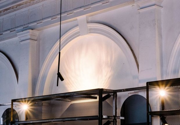 Interior architectural detail featuring ornate white plaster walls with large recessed arched niches, layered mouldings, and a decorative cornice. A contemporary black metal mesh mezzanine or balustrade crosses the lower portion of the frame, with suspended black lighting and warm spotlights creating a dramatic contrast between historic detailing and industrial elements.