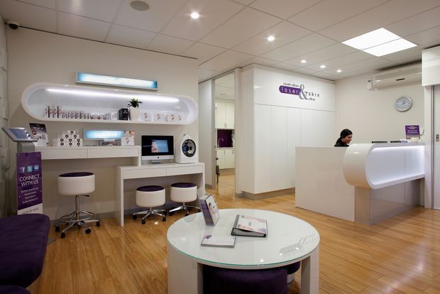 A bright, polished optical retail and consultation reception space with white built-in display cabinetry, illuminated product niches, a curved white reception desk, stools, a round glass-topped table, and warm timber flooring. The space has a suspended acoustic ceiling with recessed lighting and a clean, contemporary commercial fit-out.