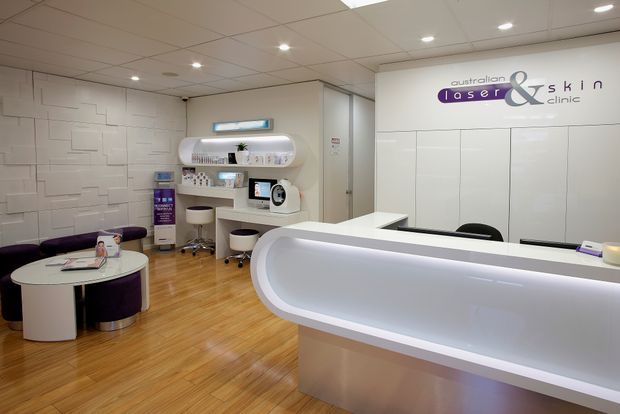 A bright, contemporary laser and skincare clinic reception area with a large curved white reception desk in the foreground. The space features pale timber flooring, white geometric wall panelling, integrated display shelving, modern salon equipment, and a compact waiting area with rounded purple seating and a small circular table. Recessed ceiling lighting creates a clean, professional atmosphere.