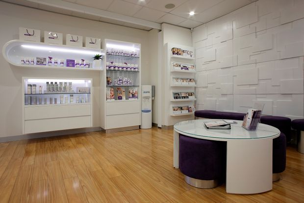 Bright, modern retail consultation and display space with white built-in product shelving, a textured geometric feature wall, polished timber flooring, and a round glass-topped consultation table surrounded by deep purple upholstered seating.