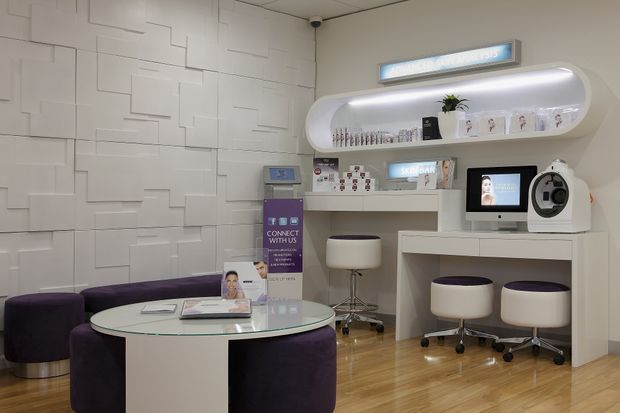 A compact, polished consultation and reception area with a curved white built-in shelving and counter unit, integrated computer workstation, display materials, stools, and a round glass-topped table surrounded by deep purple upholstered seating. The space features light timber flooring, white geometric textured wall panelling, and cool integrated lighting.