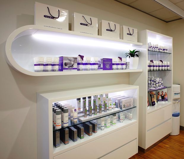 A bright retail display area, likely in a beauty or skincare clinic, featuring illuminated white built-in shelving and glass-front display cabinets stocked with packaged products. Branded shopping bags sit above the curved feature shelf, with a potted plant, pale walls, and timber flooring.