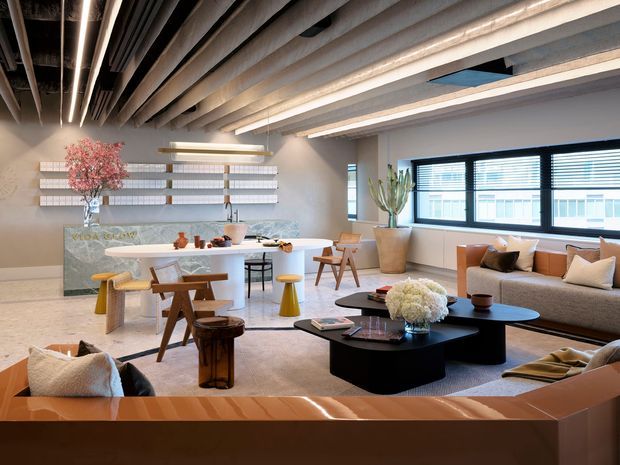 A sophisticated open-plan living and dining area with a compact kitchen island, sculptural furniture, a large sectional sofa, and a black coffee table. The space features exposed linear ceiling beams and services, expansive horizontal windows with blinds, pale textured walls, a light neutral carpet, and warm timber and brass accents. A dramatic floral arrangement and indoor plants add softness to the industrial-inspired interior.