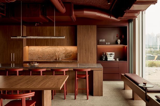 A warm, richly toned contemporary kitchen and dining area with dark timber cabinetry, a stone island and backsplash, red bar stools, and exposed ceiling ductwork. A built-in coffee and drinks station sits beside the kitchen, while a long dining table and window bench occupy the foreground. Full-height glazing provides an urban outlook.