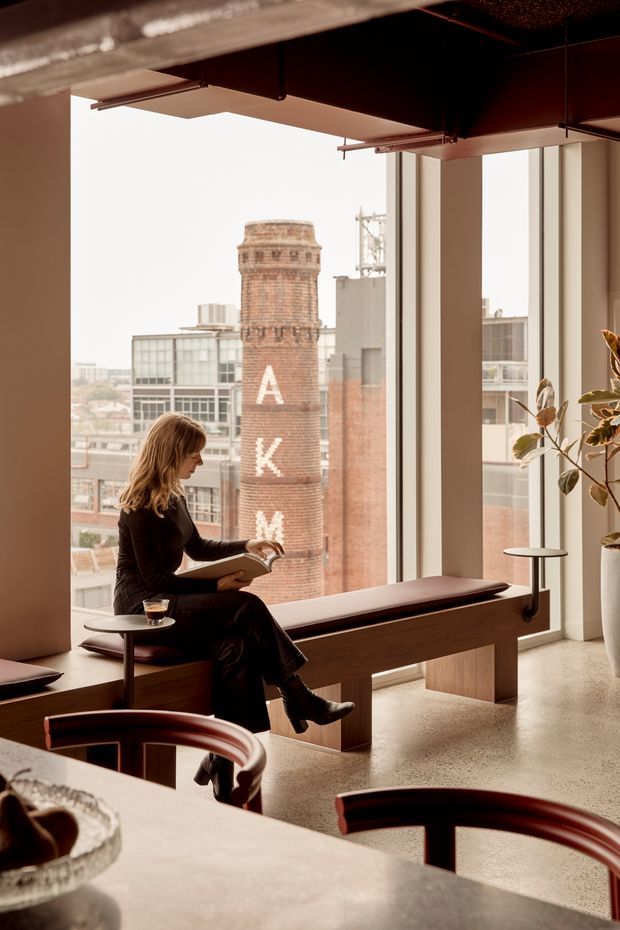 A warm, refined commercial interior with expansive floor-to-ceiling windows overlooking an urban cityscape. A woman reads while seated on a long built-in timber bench beside the glazing, with small round side tables, curved dark-wood seating in the foreground, a large stone or concrete counter, and a tall indoor plant. The space has exposed dark ceiling services, polished concrete or terrazzo flooring, and strong architectural framing around the windows.