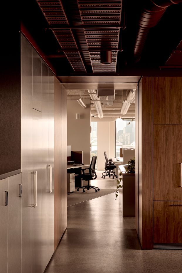 A dimly lit commercial office interior viewed from a corridor or entry passage. The foreground is framed by tall built-in cabinetry in warm wood on the right and pale metallic or light-toned cabinetry on the left. Beyond, an open-plan workspace contains desks, computer monitors, office chairs, and indoor plants, with city buildings visible through large windows. The exposed ceiling features dark perforated metal panels, visible ductwork, and industrial services, creating a refined industrial atmosphere.