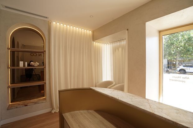 A warm, softly lit reception or waiting area with a curved built-in shelving niche, illuminated arch, vertical sheer curtains, a large wall mirror, and a marble-look reception counter with integrated timber seating. Timber flooring and neutral plastered walls create a refined, calm interior.