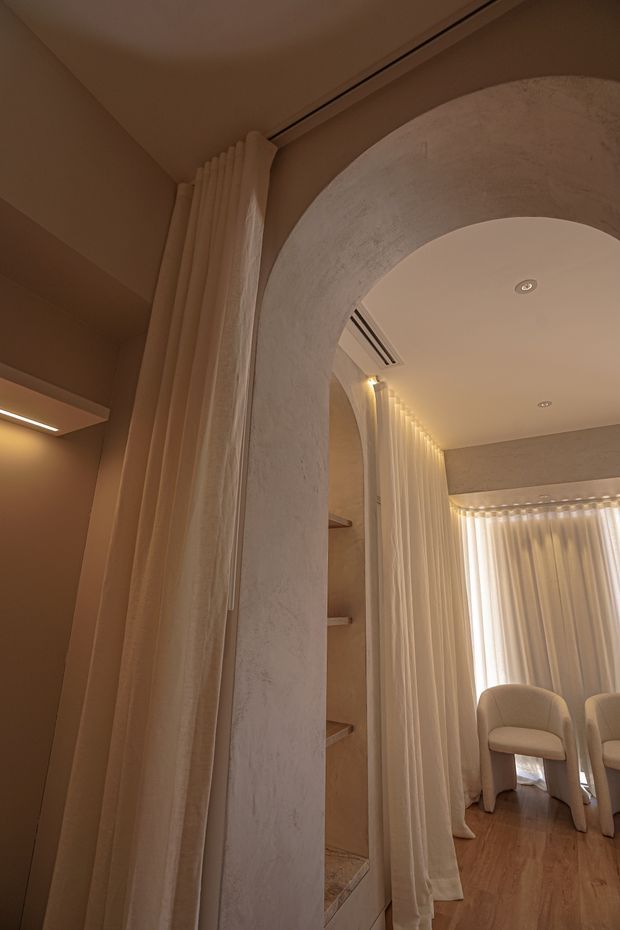 Warm, softly lit interior featuring a large rounded plaster archway, layered full-height cream curtains, recessed shelving, and pale timber flooring. Through the arch is a refined seating area with upholstered chairs and sheer curtains, creating an intimate, softly textured atmosphere.