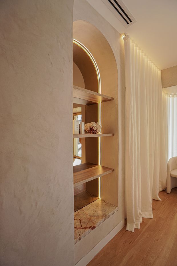 Warm, refined interior detail featuring a built-in arched display niche with illuminated curved edging, timber shelves, and a veined stone base. Textured beige plaster walls, sheer floor-length curtains, timber flooring, and soft concealed lighting create an elegant, softly layered atmosphere. A partial upholstered chair is visible at right.