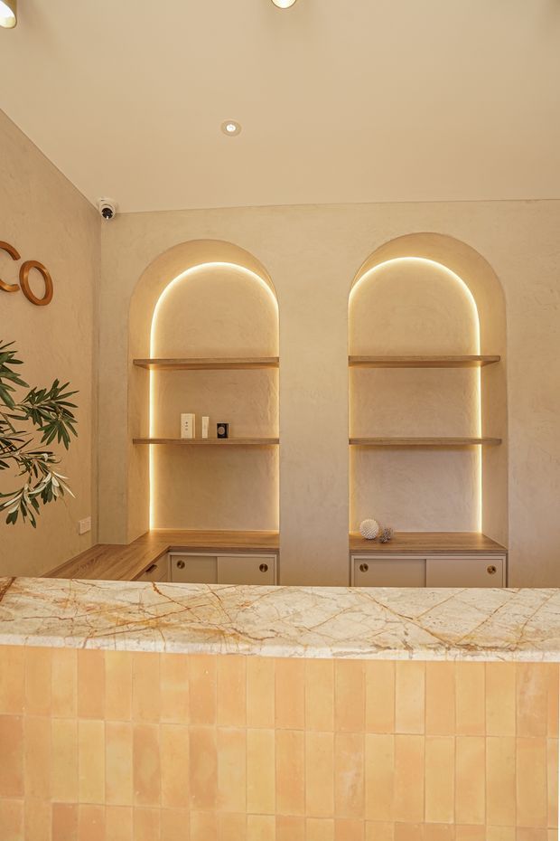 Warm, intimate interior view of a refined reception or serving counter, featuring a tiled terracotta-toned front, a veined natural stone benchtop, and illuminated arched built-in display niches with timber shelves and drawers. Plastered walls, recessed ceiling lights, and subtle decorative objects create a softly atmospheric, Mediterranean-inspired setting.