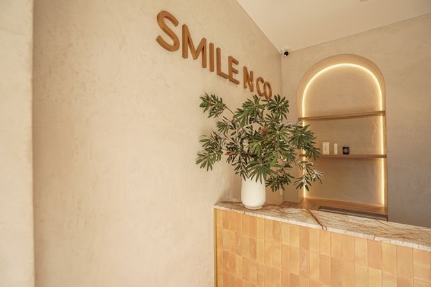Warm, softly lit reception counter in a dental or medical clinic, featuring a textured beige plaster wall, large raised wooden “SMILE NO...” lettering, a tiled front counter with a light stone benchtop, and a curved recessed display niche with integrated LED lighting and timber shelves. A potted tree adds a natural accent.