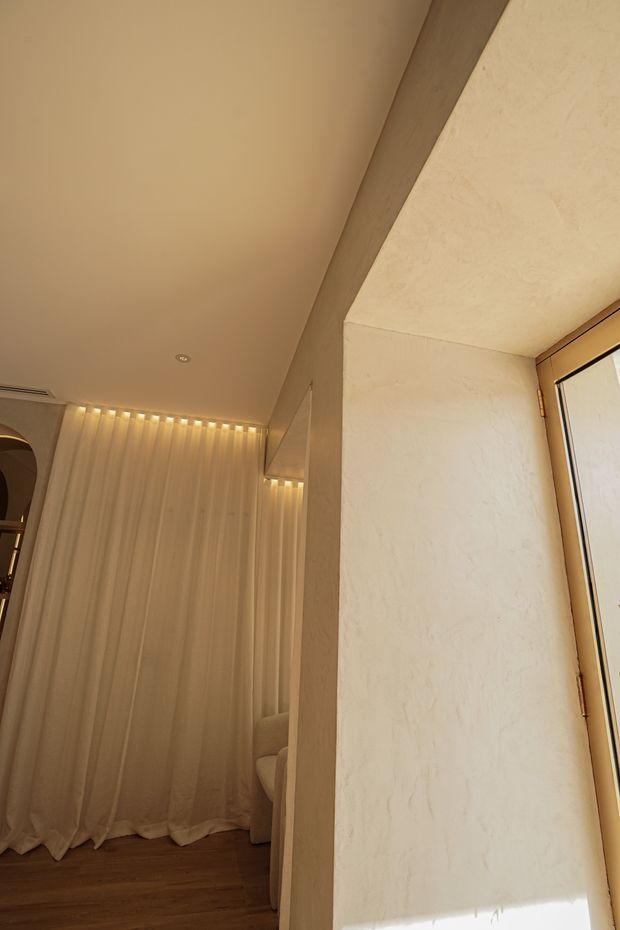 Warm, softly lit interior detail showing a textured off-white plaster wall, timber-toned ceiling beam and flooring, full-height sheer curtains with concealed linear lighting, and a partial upholstered seating area. A glazed timber-framed opening is visible on the right. The space has a refined, earthy and minimalist feel.