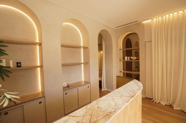 Warm, softly lit contemporary interior featuring a curved marble-clad island or counter in the foreground, arched recessed shelving with integrated LED lighting and lower cabinetry, timber flooring, and full-height cream curtains. The space has a refined, organic feel with rounded architectural detailing and brass-toned joinery.