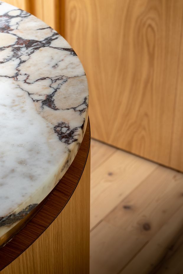 Close-up detail of a rounded white natural stone benchtop with pronounced dark veining, set above curved dark timber edging and fluted or finely grooved timber cabinetry. Warm timber wall panelling and a light timber floor create a refined, tactile interior joinery composition.