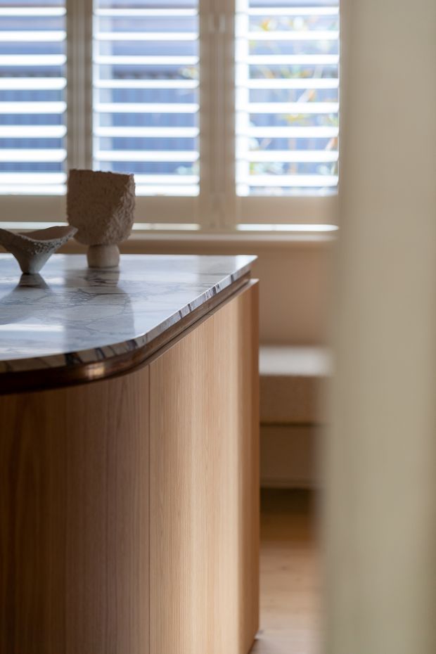 Close-up interior view of a refined timber kitchen or built-in cabinetry, featuring vertically grained light-to-mid wood doors, a rounded polished stone benchtop with a dark edge detail, and warm neutral walls. White plantation shutters provide filtered daylight in the background, while minimal sculptural objects sit on the benchtop.