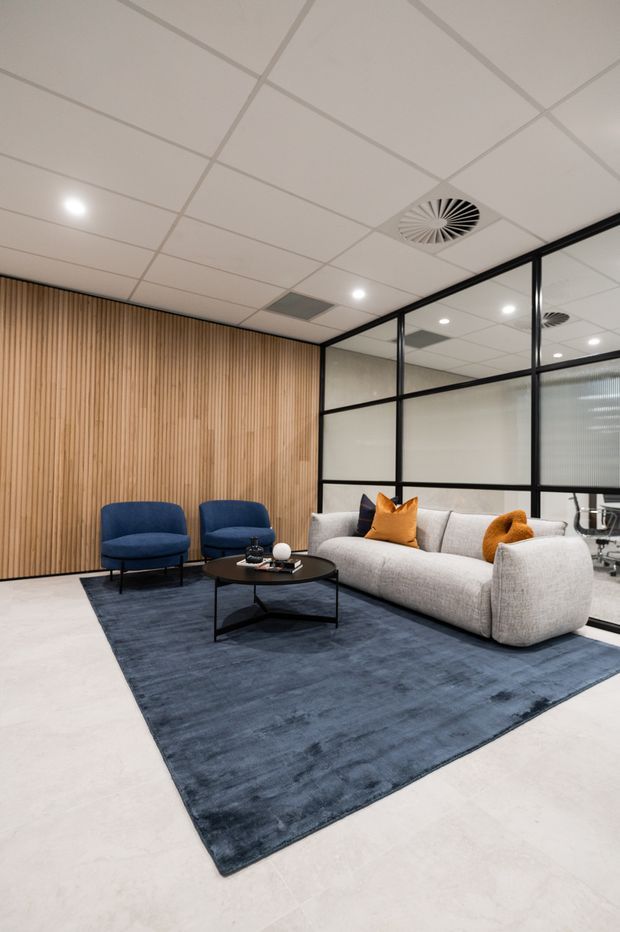 A contemporary office lounge or waiting area with a large light-grey sectional sofa, two navy armchairs, a black coffee table, and a dark blue rug. The room features a warm vertical timber slat feature wall, black-framed frosted and clear glass partitions, a white suspended acoustic-tile ceiling with recessed lighting and circular air vents, and a glimpse of office workstations beyond the glazing.