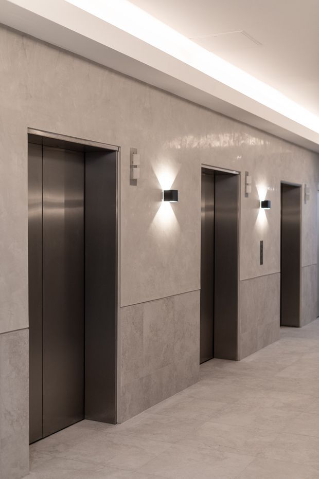A refined elevator lobby with three dark metal elevator doors set into a continuous pale grey textured wall finish. Minimal black-and-white wall sconces illuminate the corridor, while large-format pale flooring and a recessed linear ceiling detail create a clean, understated commercial interior.