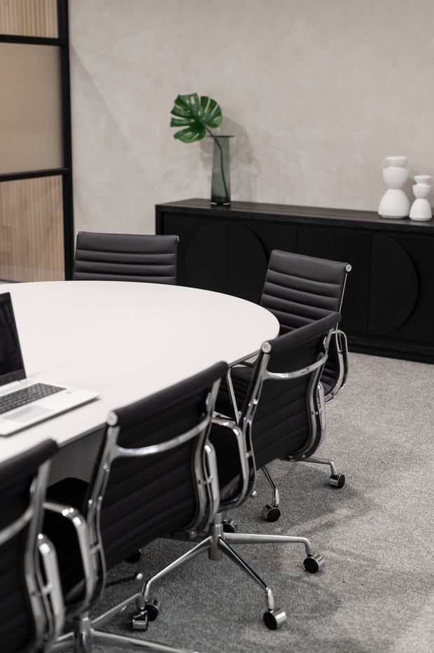 A refined contemporary meeting room with a large round white table, black leather office chairs with polished metal frames, and a black curved-door credenza. The room features a textured warm off-white wall, grey carpet, black-framed fluted-glass partitioning, a simple leafy arrangement, and sculptural white vases.