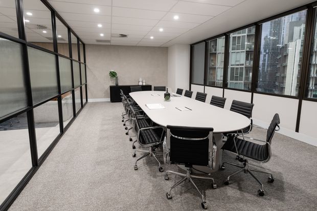 A contemporary commercial meeting room with a long white oval conference table surrounded by black office chairs. The room has grey carpet, white acoustic ceiling tiles with recessed downlights, black-framed glazed partitions, large windows overlooking urban buildings, and a minimal black storage cabinet with a small plant.