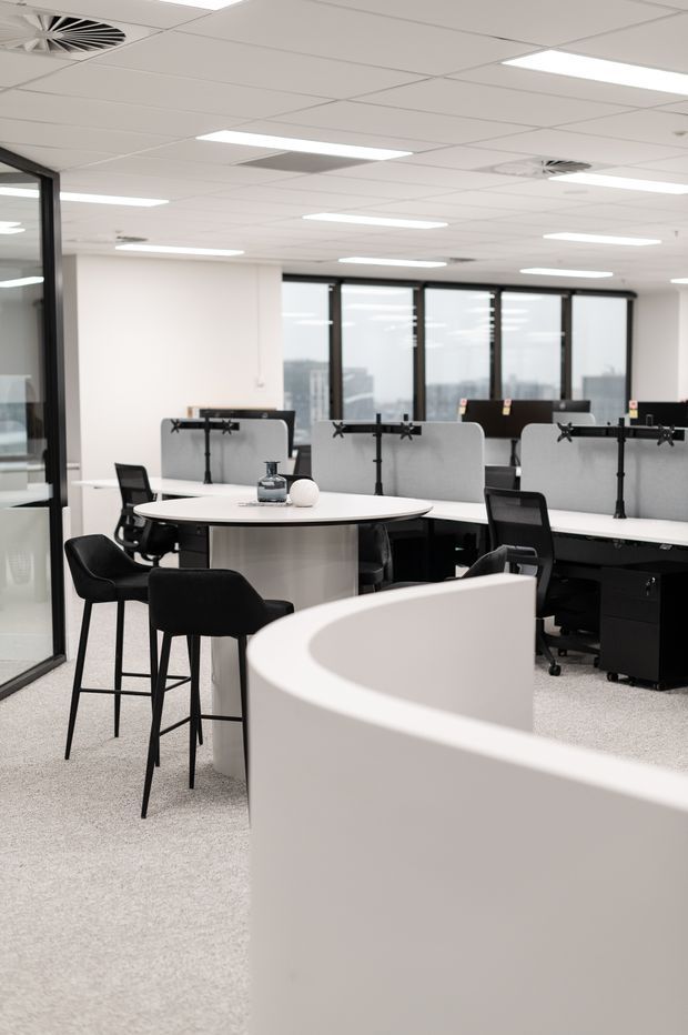 A bright, contemporary open-plan office interior with shared workstations, black office chairs, white curved desks, acoustic privacy screens, and a small round meeting table with bar-height seating. The space has a suspended white grid ceiling with recessed linear lighting, dark-framed glass partitions, and large windows overlooking an urban setting.