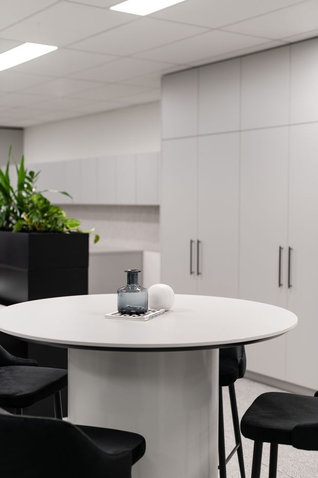 A minimalist contemporary office or commercial interior with a round white table and central pedestal, surrounded by black upholstered chairs. White full-height built-in cabinetry lines the rear wall, with a suspended acoustic tile ceiling, recessed fluorescent-style lighting, and a large black planter containing greenery. The palette is predominantly white, grey, and black with a clean, refined appearance.