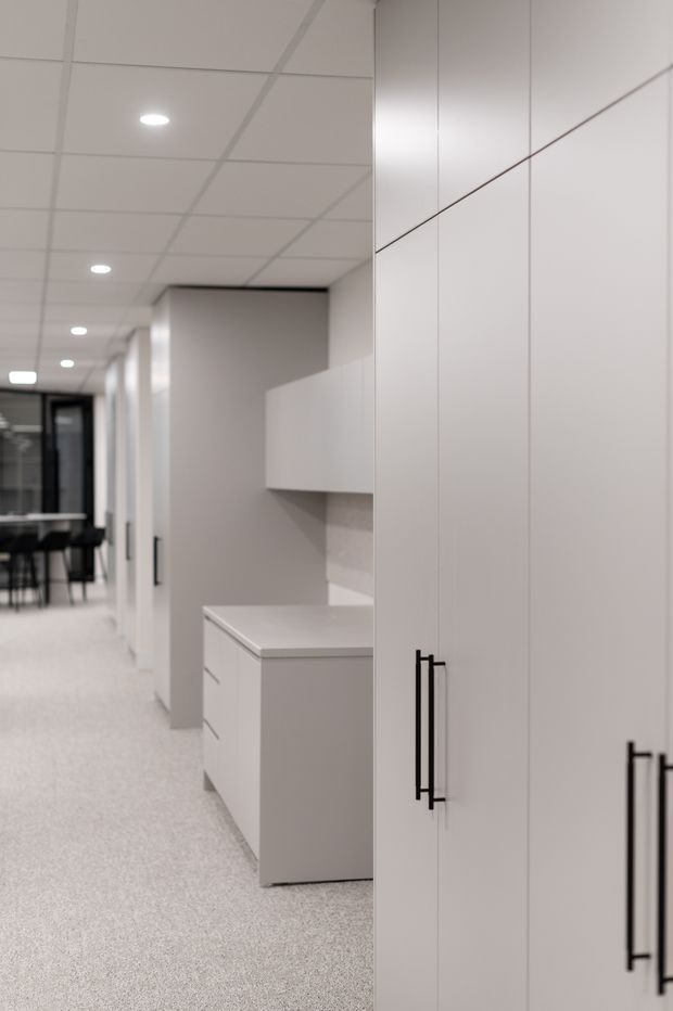 A bright, minimalist commercial interior showing a long corridor lined with tall white built-in storage cabinetry and black vertical handles. A compact kitchenette or workroom joinery area is visible midway down the corridor, with a communal table and chairs at the far end. The space has a suspended white grid ceiling with recessed downlights, pale grey walls, and a light speckled flooring finish.