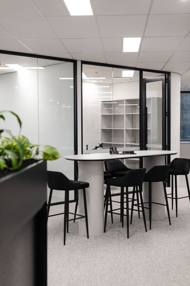 A contemporary commercial office breakout area with a tall white communal table, black bar-height chairs, and a black-framed glass meeting room enclosure. The space has white acoustic ceiling tiles with recessed fluorescent lighting, light grey carpet, white walls, and built-in open shelving visible through the glass. A large black planter with greenery occupies the foreground.