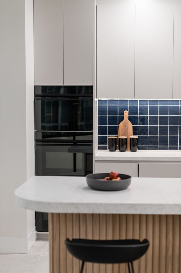 Contemporary kitchen featuring tall integrated black appliances, pale cabinetry, a dark blue rectangular tile splashback, a light stone island benchtop, and a fluted timber island front with a black bar stool. The kitchen is clean-lined and softly lit, with restrained neutral tones and dark accents.