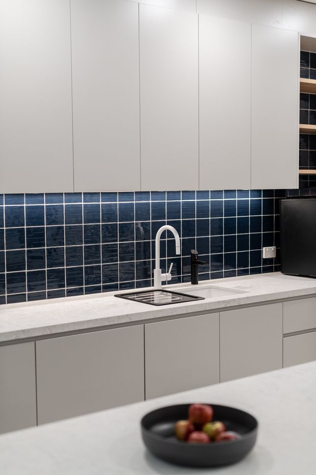 A compact contemporary kitchen with flat-front light grey cabinetry, a white stone-look benchtop, and a striking dark navy-blue square tile splashback. The kitchen includes an undermount sink, curved white tap, contrasting black mixer, integrated storage, and a foreground island or peninsula with a black fruit bowl.