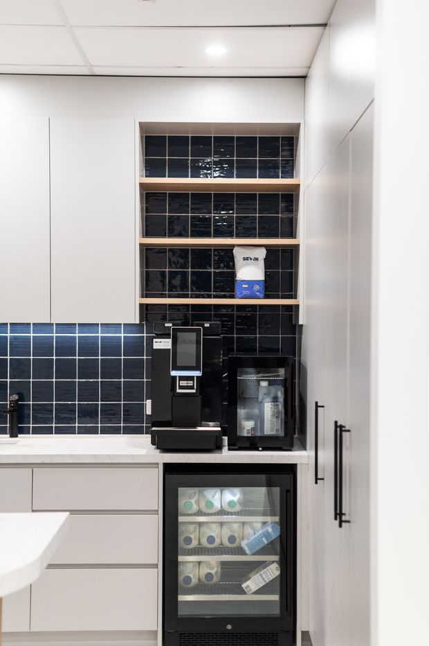 Compact modern kitchenette or office refreshment station with white flat-panel cabinetry, a pale stone benchtop, glossy dark blue-black square tiled splashback, open timber shelves, coffee machine, under-bench drinks refrigerator, and tall storage cabinets. The clean, functional fit-out has bright recessed lighting and strong black-and-white contrast.