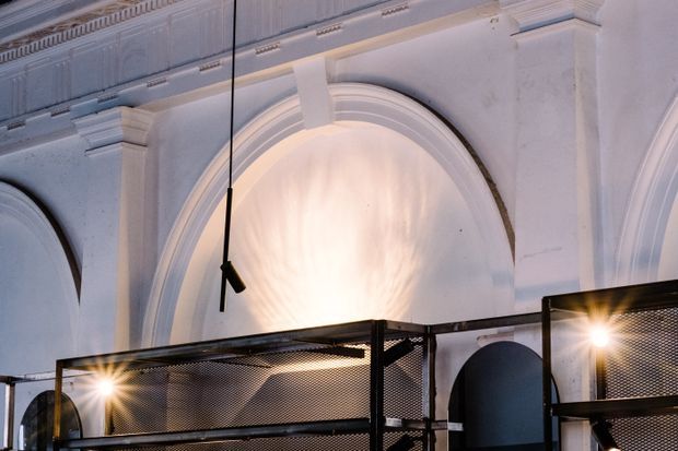 Interior architectural detail featuring ornate white plaster walls with large rounded arches, layered mouldings, pilasters, and decorative cornices. A contemporary black metal mezzanine or balustrade with perforated mesh panels crosses the lower foreground, with modern suspended lighting creating a strong contrast between historic detailing and industrial elements.