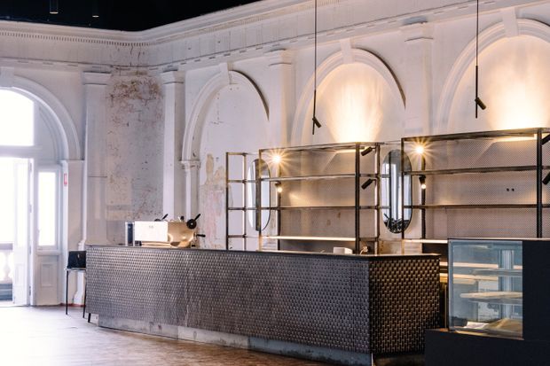 A dramatic commercial interior, likely a café or hospitality counter, set within a large heritage-style room. The space features ornate white plaster wall arches and mouldings, weathered wall surfaces, a long dark tiled service counter, black metal and mesh display shelving, pendant spotlights, and a glass-fronted refrigerated display case. Bright daylight enters through tall arched openings on the left, contrasting with the dark ceiling and industrial fit-out.