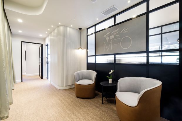 Contemporary reception or waiting area with two rounded upholstered lounge chairs and a small black side table, positioned beside a black-framed glazed partition. The space features a warm beige carpet, white walls and ceiling, a curved vertically ribbed feature wall, recessed lighting, and a subtle MFO graphic on the glazing. An open doorway leads to an adjacent corridor or room.